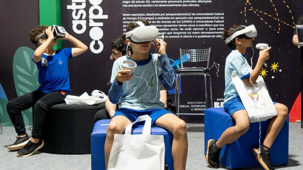 foto de estudantes crianças sentados em puffs de cor azul e preto em formato de circulo, usando óculos de realidade 3D durante a 21 semana nacional de ciência e tecnologia