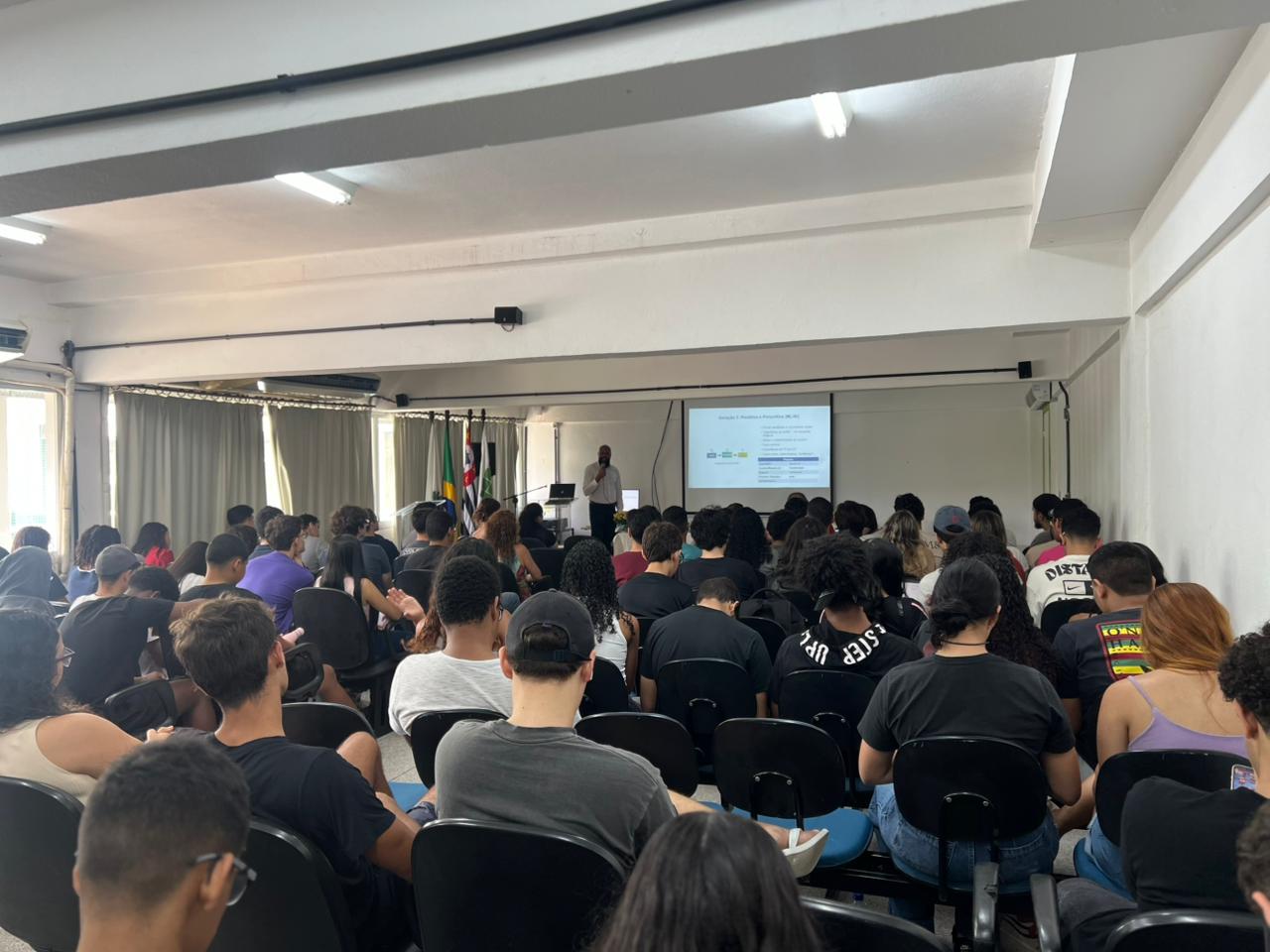 imagem de um auditório fechado com paredes brancas e diversas pessoas sentadas em cadeiras pretas
